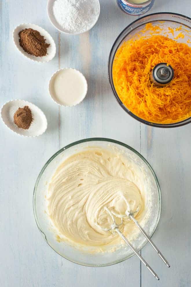 Pumpkin poke cake batter ingredients combined in a bowl using a hand mixer, surrounded by more ingredients on a countertop.