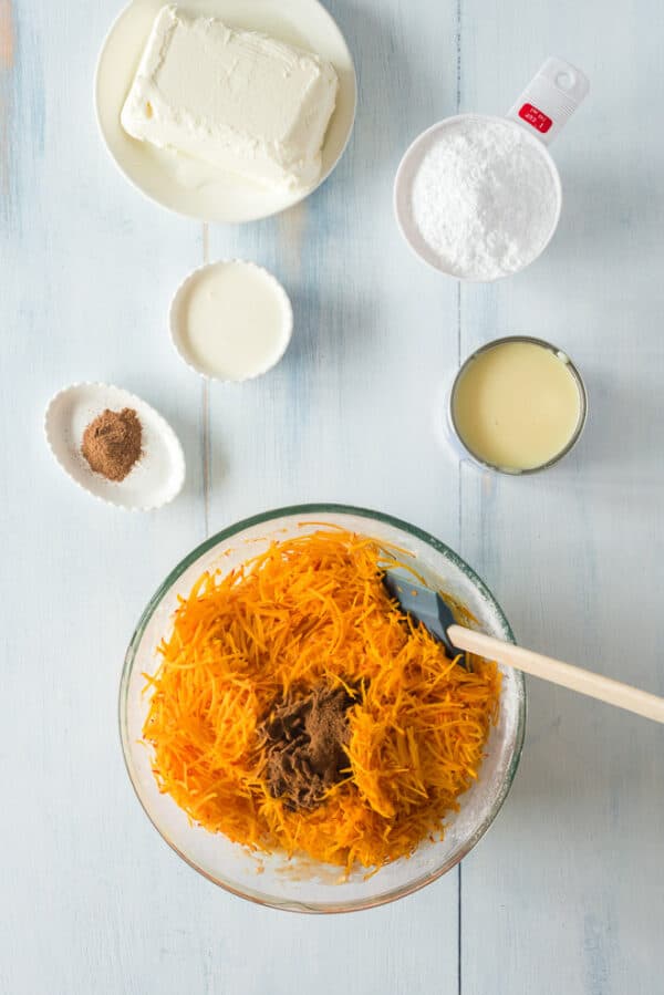 Shredded pumpkin and pumpkin spice added to a bowl of pumpkin poke cake batter, surrounded by more ingredients on a countertop.