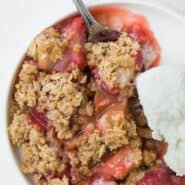 Close up view of strawberry rhubarb crisp and vanilla ice cream on a round plate, with a spoon.