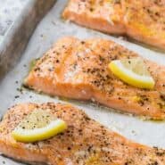 Three slow roasted salmon filets topped with lemon slices and black pepper on a parchment lined baking sheet.