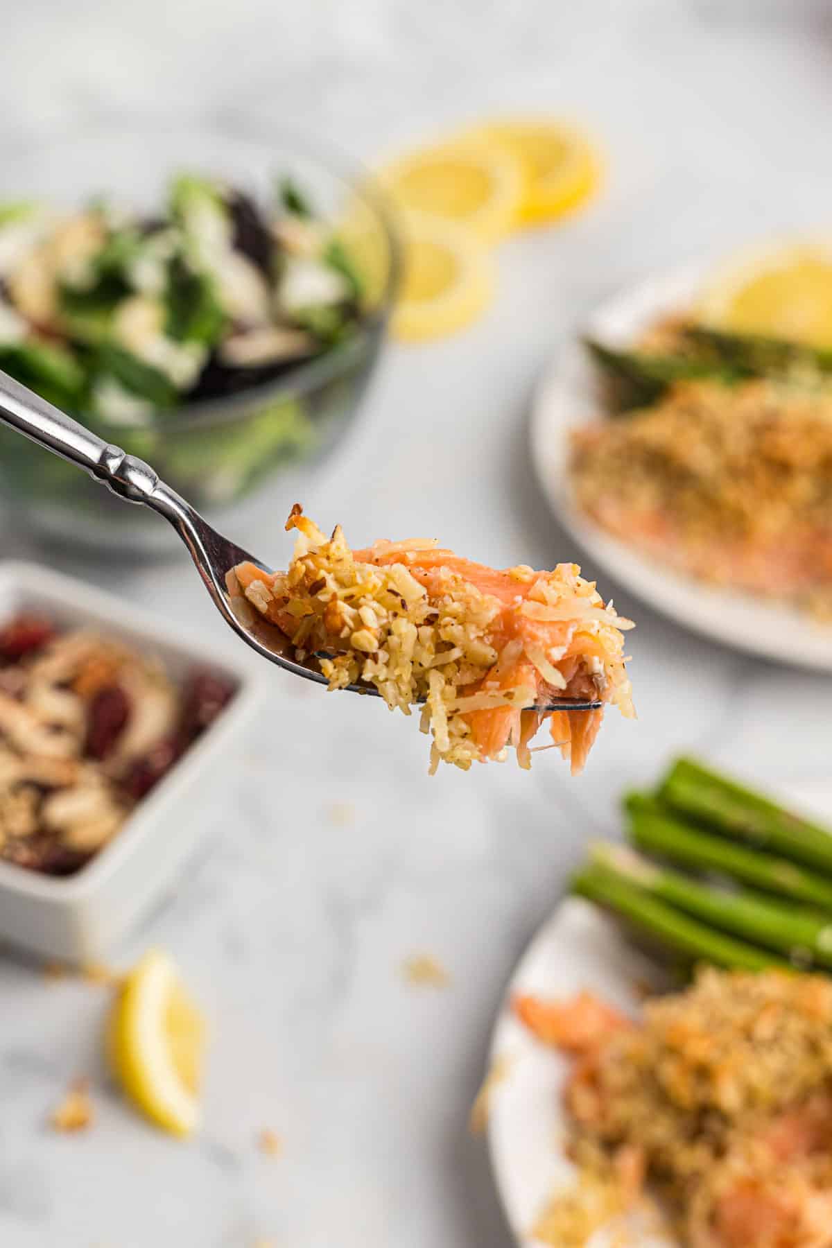 Flaky salmon on a fork, with plated entrees and asparagus in the background.