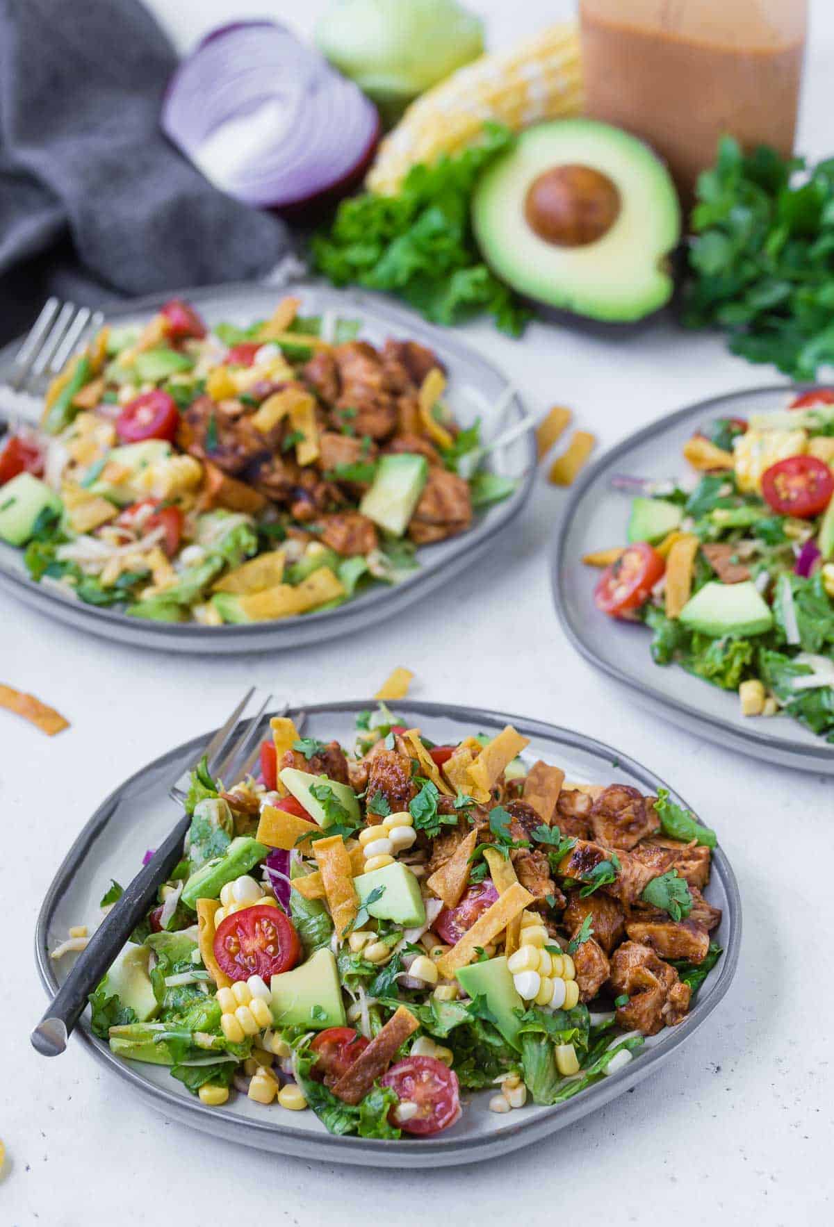 Three plates of a colorful chopped salad on a grey plates. Salad includes bbq grilled chicken, lettuce, onion, tomato, crispy tortilla strips, avocado, corn and more!