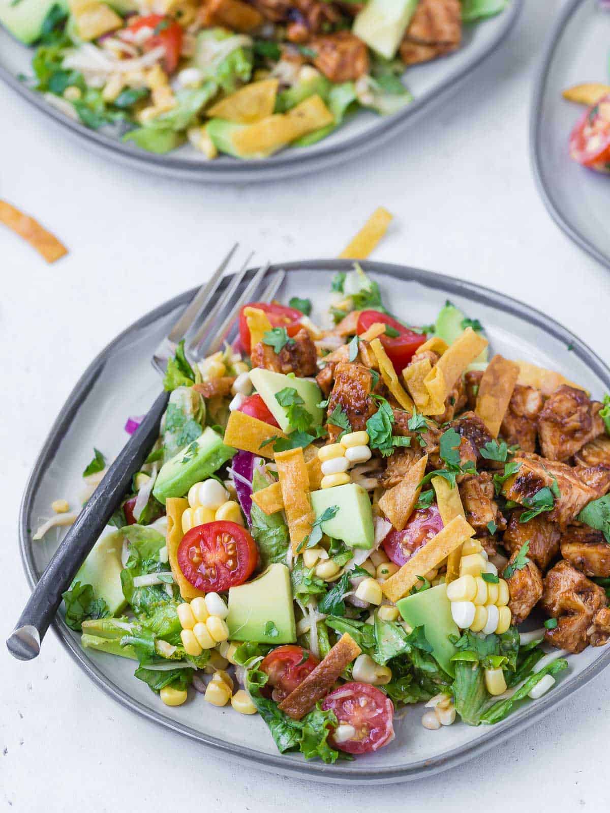 Colorful chopped salad on a grey plate. Salad includes bbq grilled chicken, lettuce, onion, tomato, crispy tortilla strips, avocado, corn and more! Whole ingredients are featured in the background of the image.