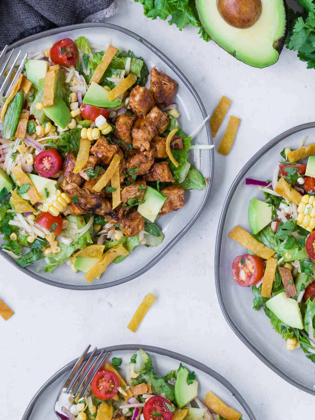 Overhead view of three plates of a colorful chopped salad on a grey plate. Salad includes bbq grilled chicken, lettuce, onion, tomato, crispy tortilla strips, avocado, corn and more!