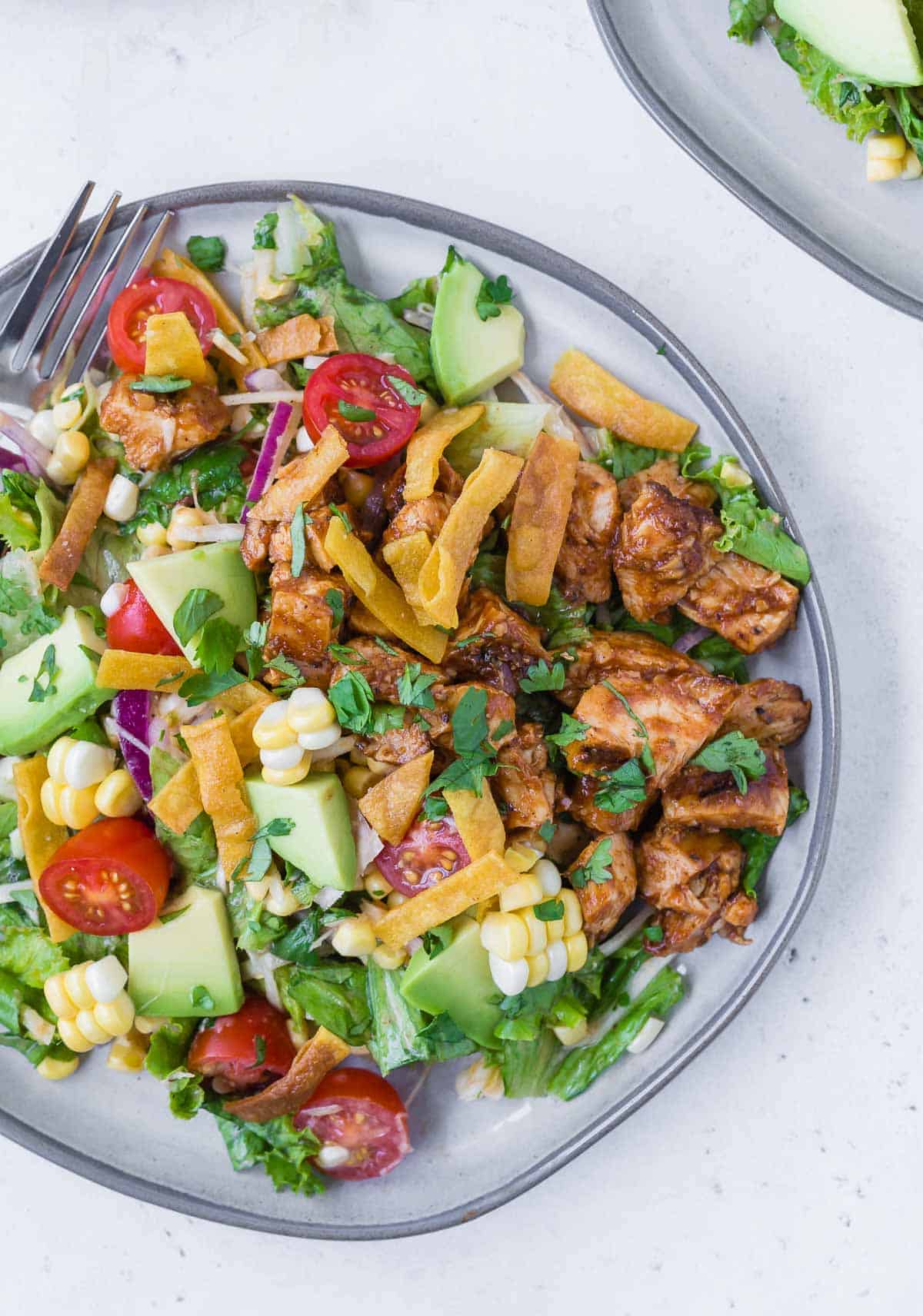 Overhead view of a colorful chopped salad on a grey plate. Salad includes bbq grilled chicken, lettuce, onion, tomato, crispy tortilla strips, avocado, corn and more!