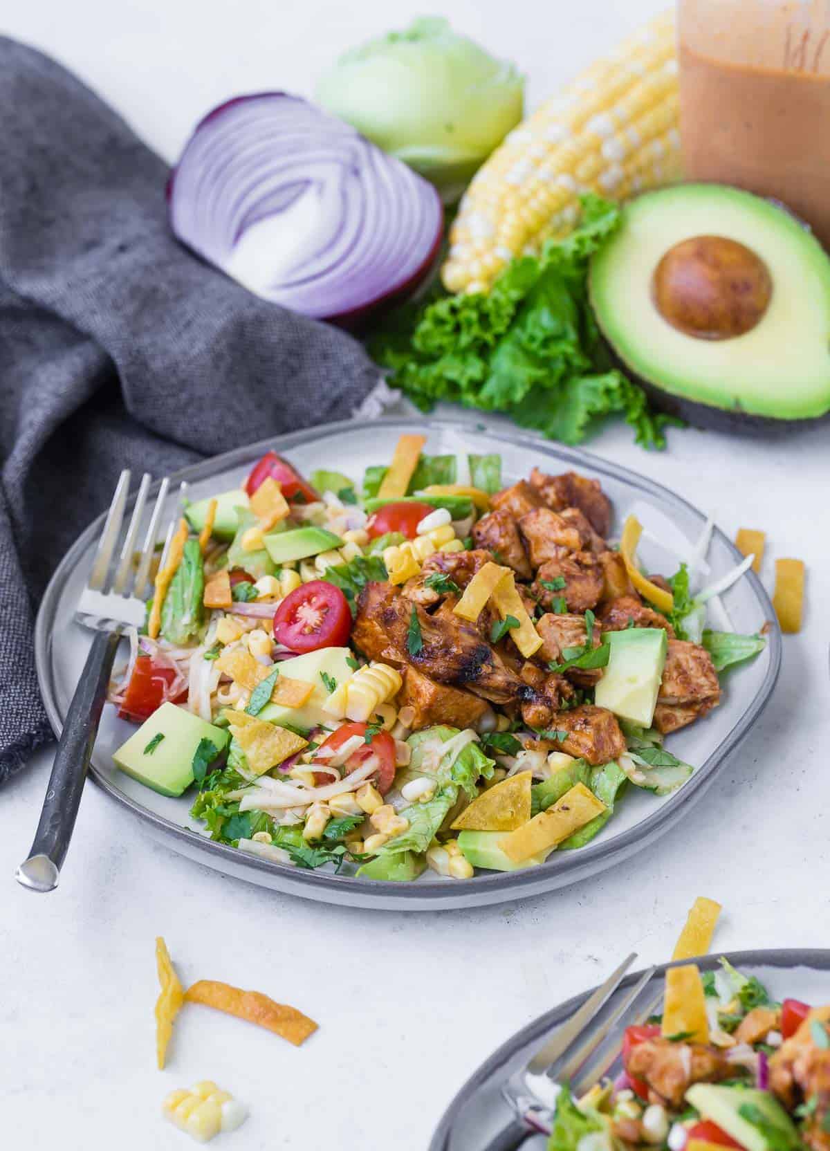 Colorful chopped salad on a grey plate. Salad includes bbq grilled chicken, lettuce, onion, tomato, crispy tortilla strips, avocado, corn and more! Whole ingredients are featured in the background of the image.