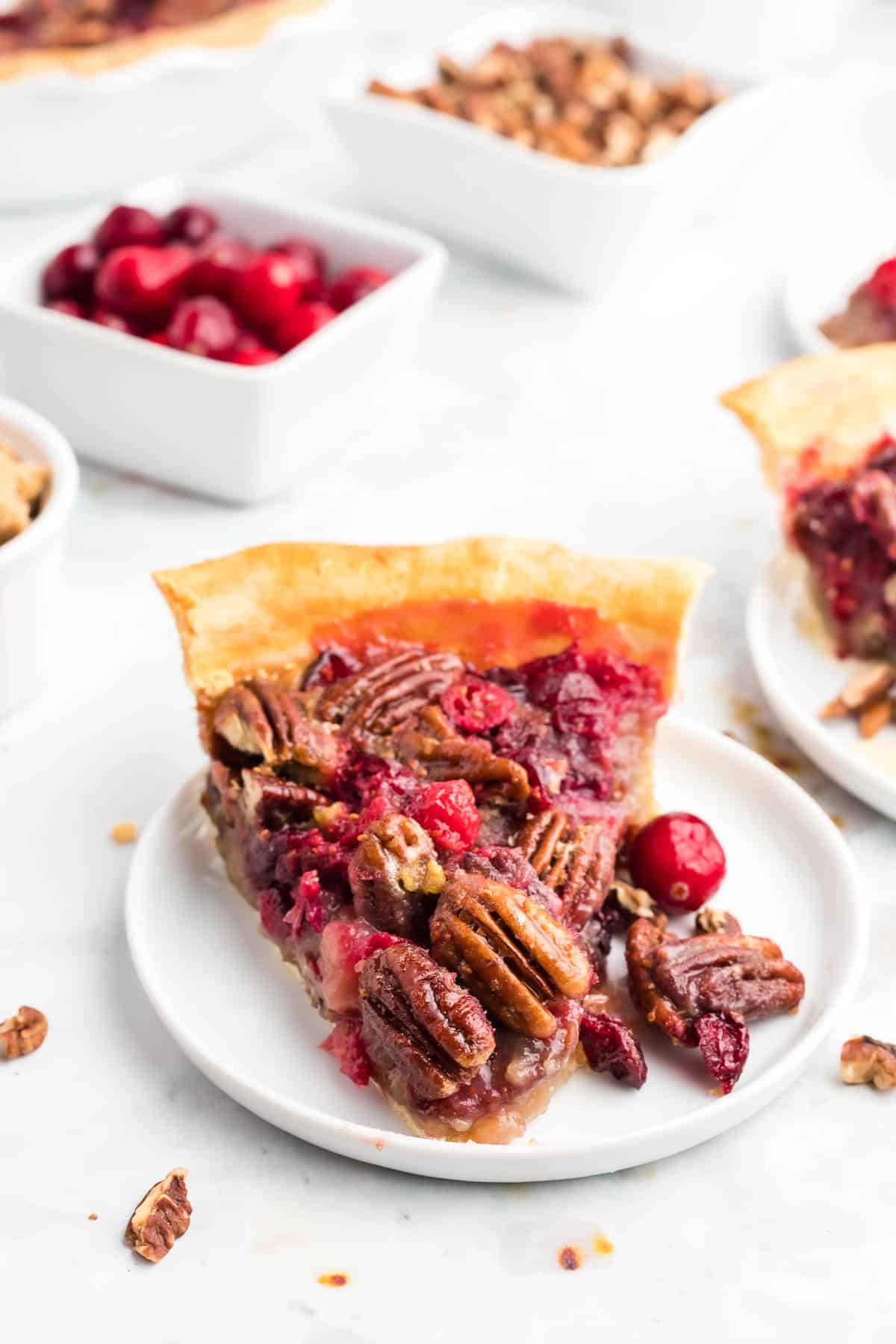 Pie slice on a white plate with cranberries and pecans scattered around.