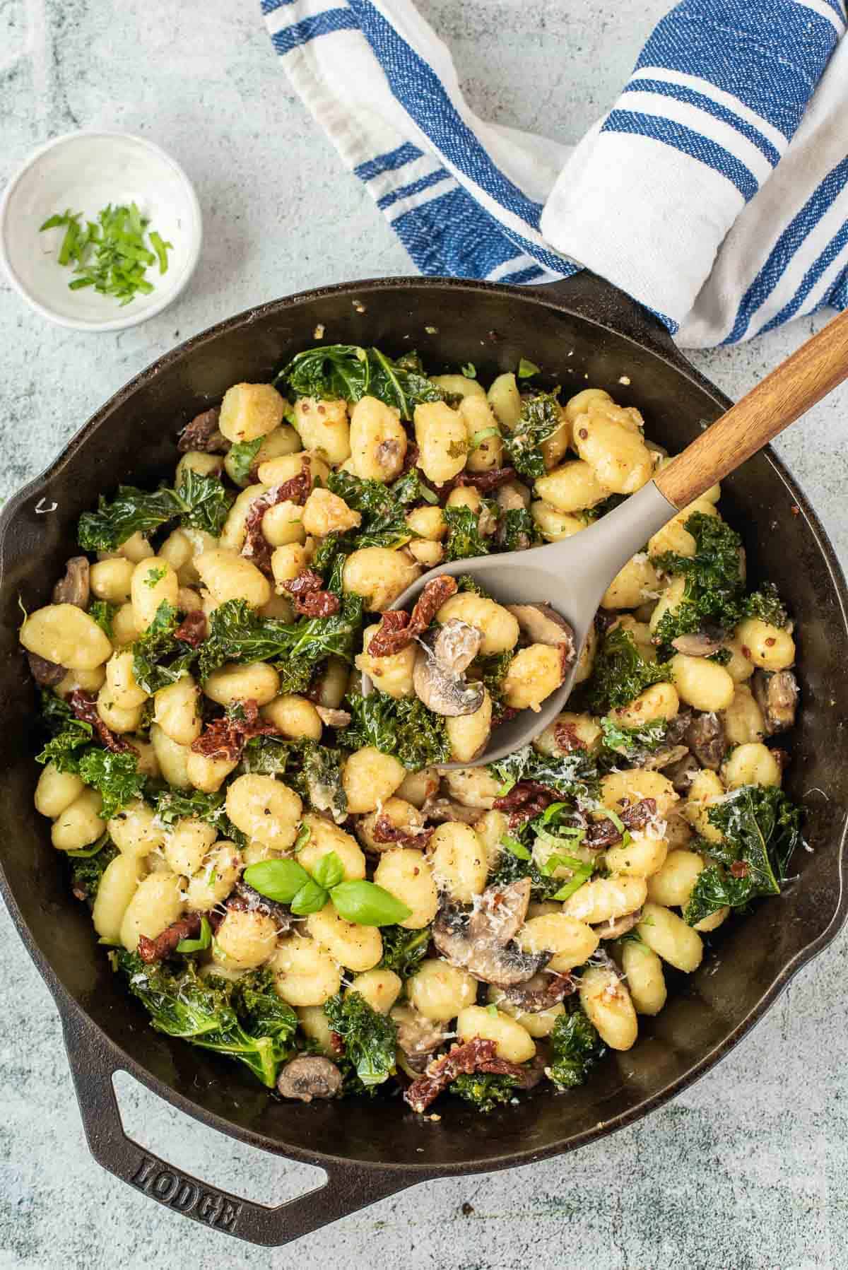 Overhead view of a black skillet filled with gnocchi with kale, sun-dried tomatoes, and mushrooms.