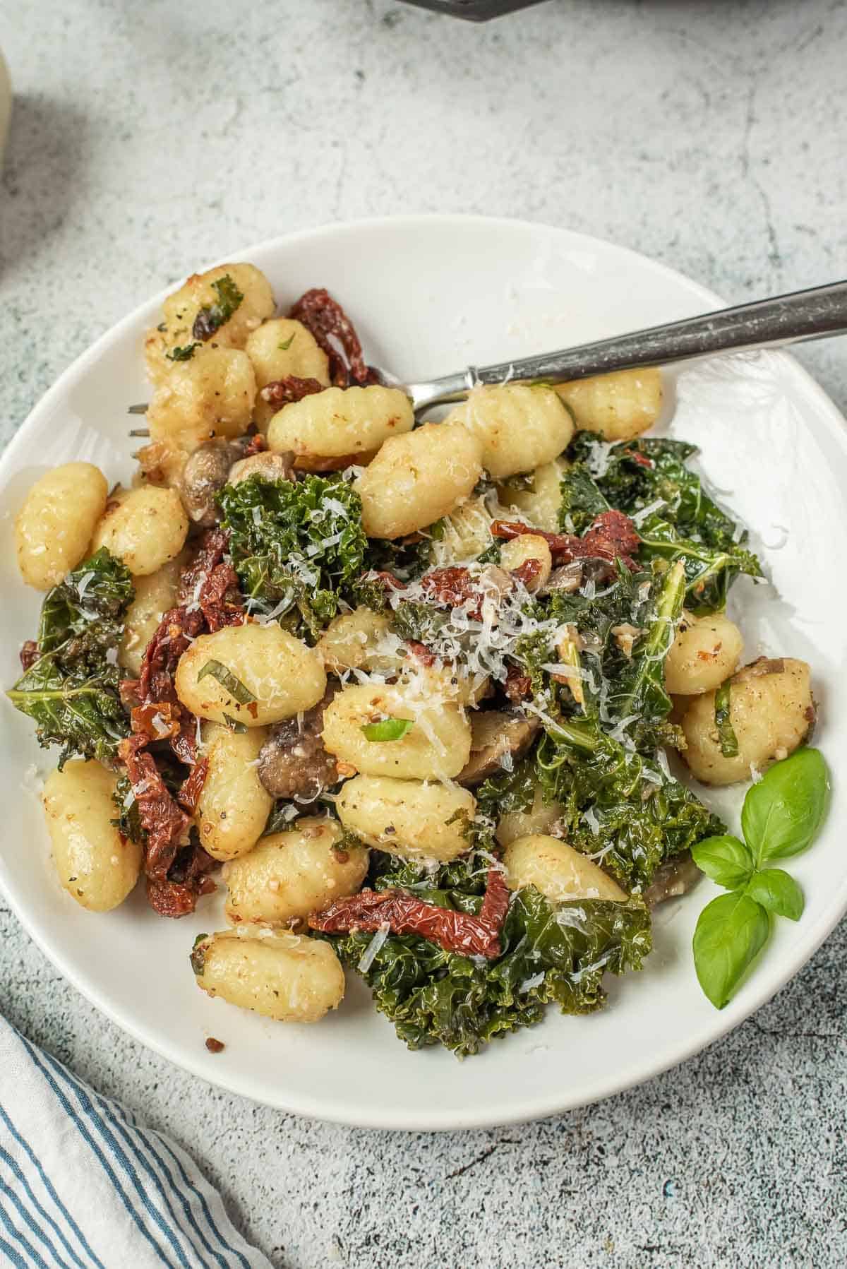 Gnocchi on a white plate with kale, mushrooms, sun-dried tomatoes, and cheese.