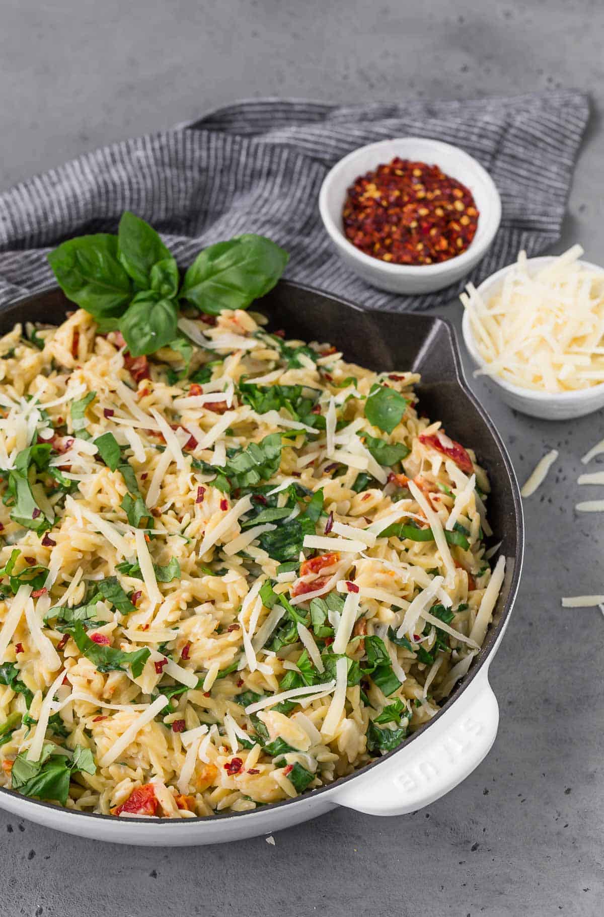 A white and black skillet on a grey surface, filled with creamy orzo with spinach, sundried tomatoes, and basil.