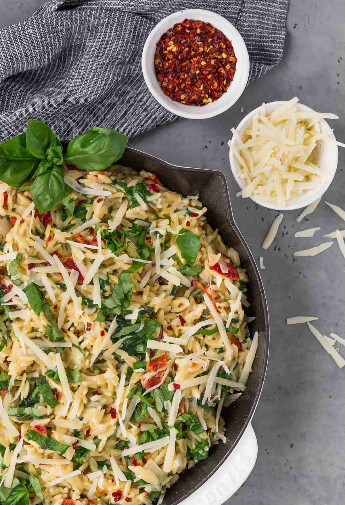 Overhead view of creamy orzo in a skillet, a bowl of parmesan cheese, and a bowl of red pepper flakes.