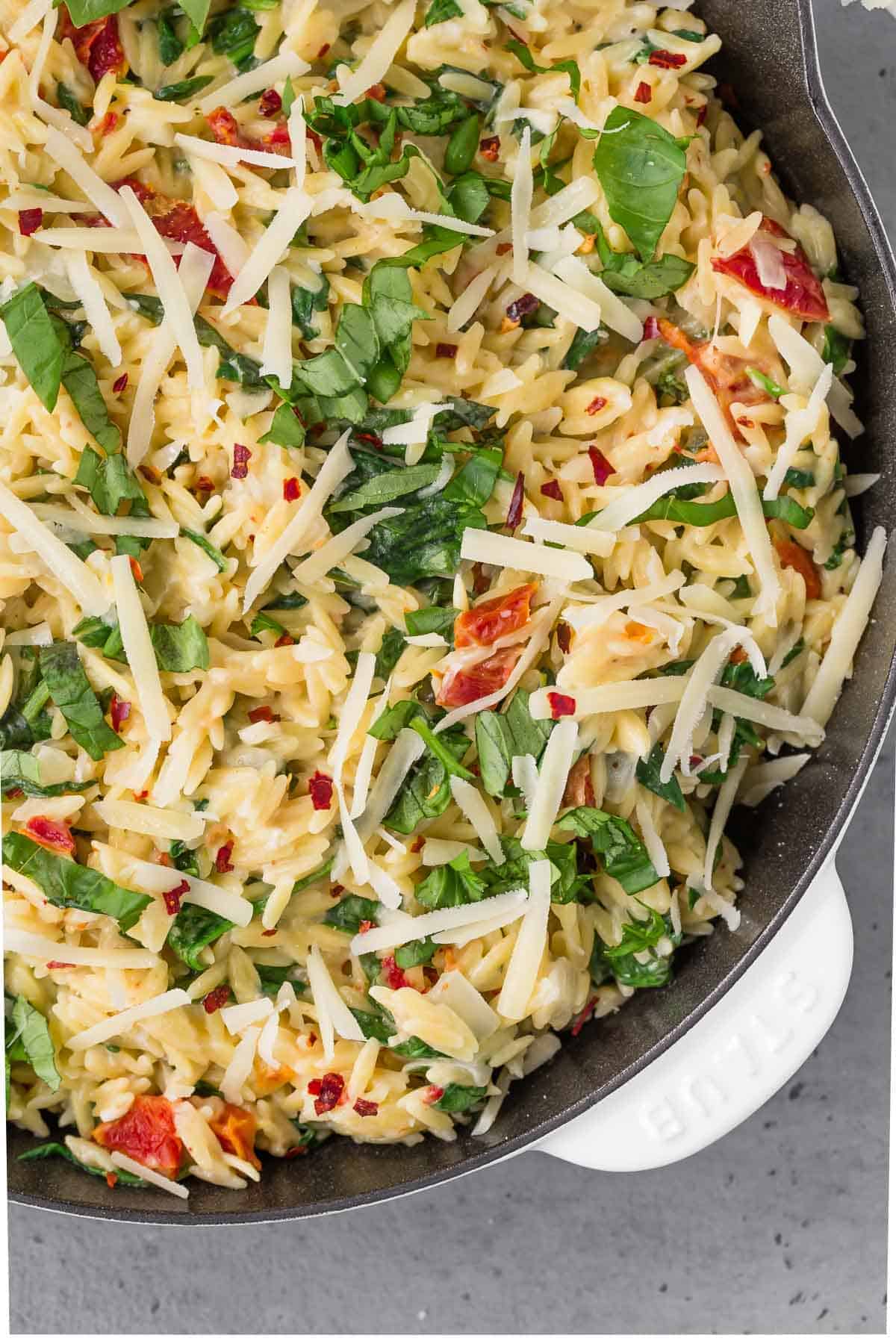 Close up overhead view of creamy orzo in a skillet with sundried tomatoes, basil, parmesan, and red pepper flakes.