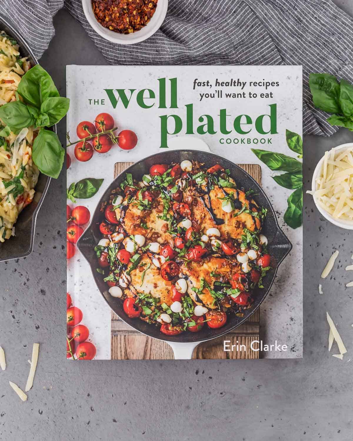 The Well Plated Cookbook on a gray surface. Also pictured are a pasta dish, a small bowl of shredded parmesan cheese, a small bowl of red pepper flakes, and fresh basil.