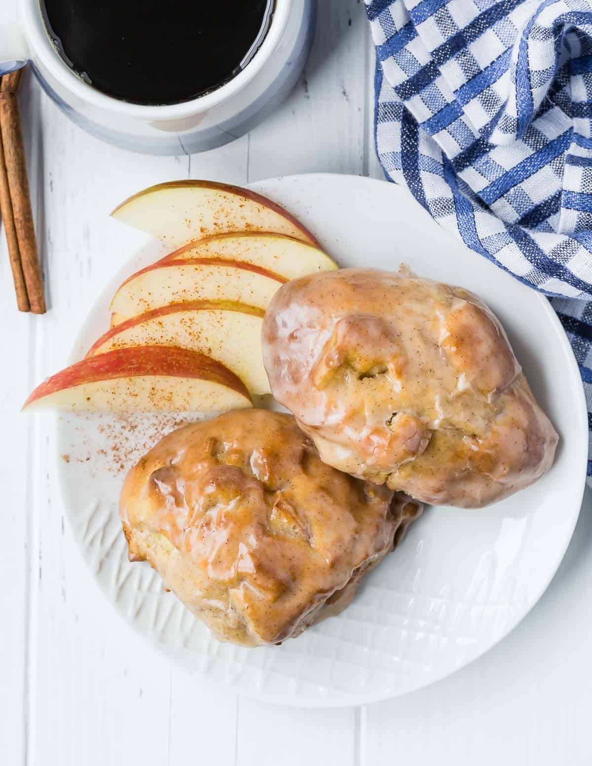 Overhead view of two glazed apple fritters on a plate, with apples. Coffee is also visible as well as a blue and white linen.