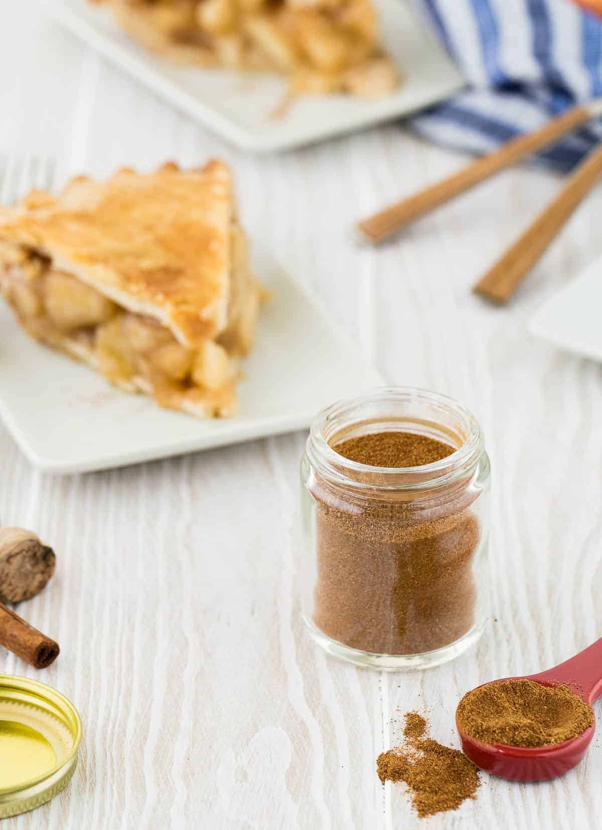 Wooden background with apple pie spice in a small jar. Also pictured are cinnamon sticks and apple pie on a white plate. 