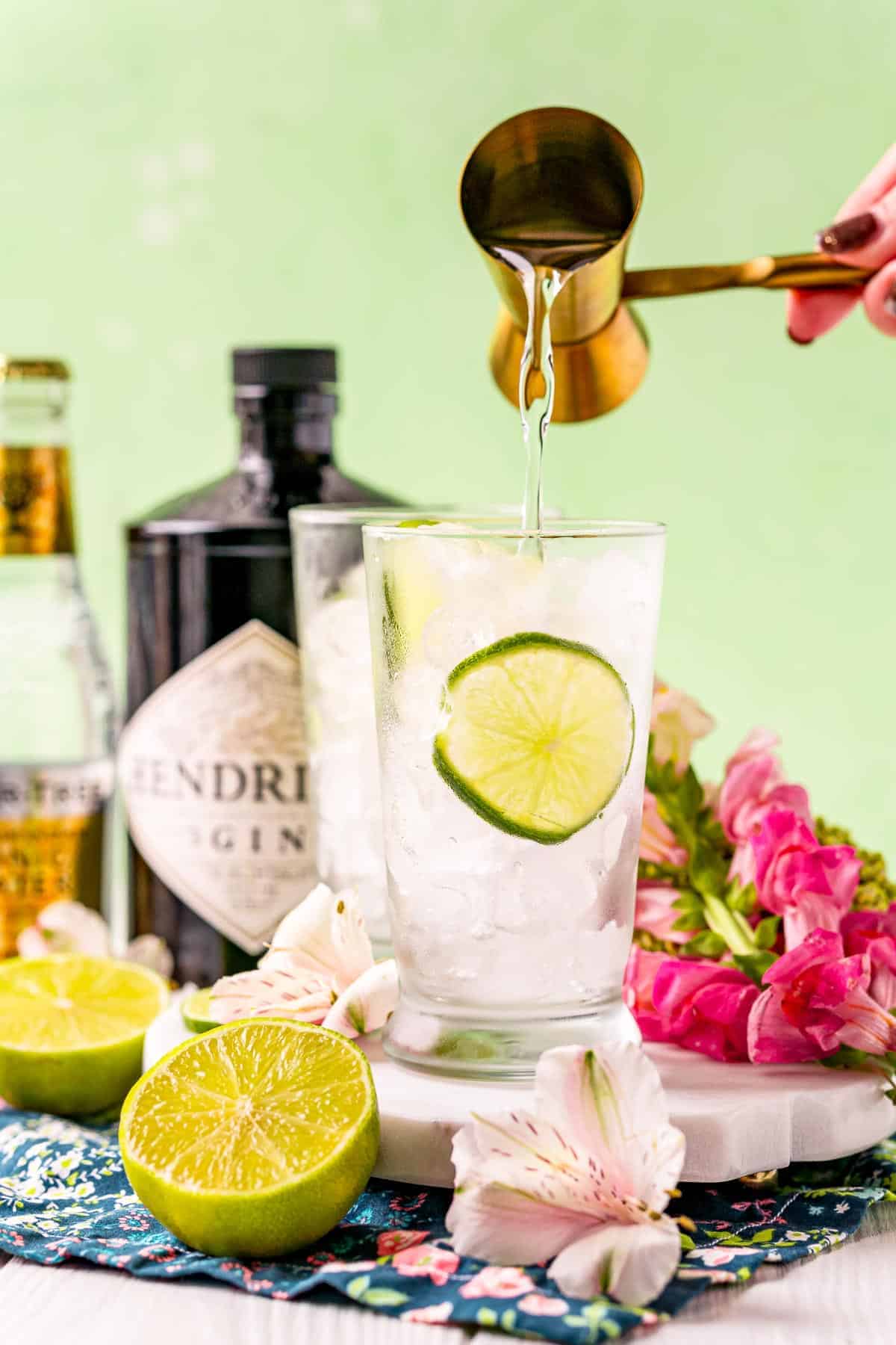 Gin being poured into a glass, a bottle of gin and a bottle of tonic pictured in the background. 