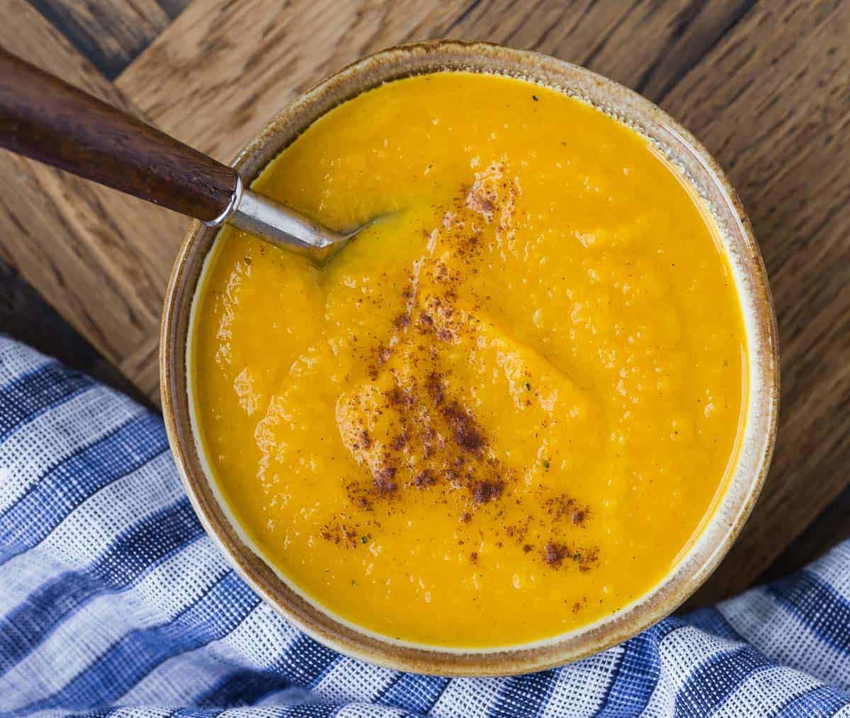 Bowl of orange colored squash bisque in a natural tone bowl with a wooden handled spoon.