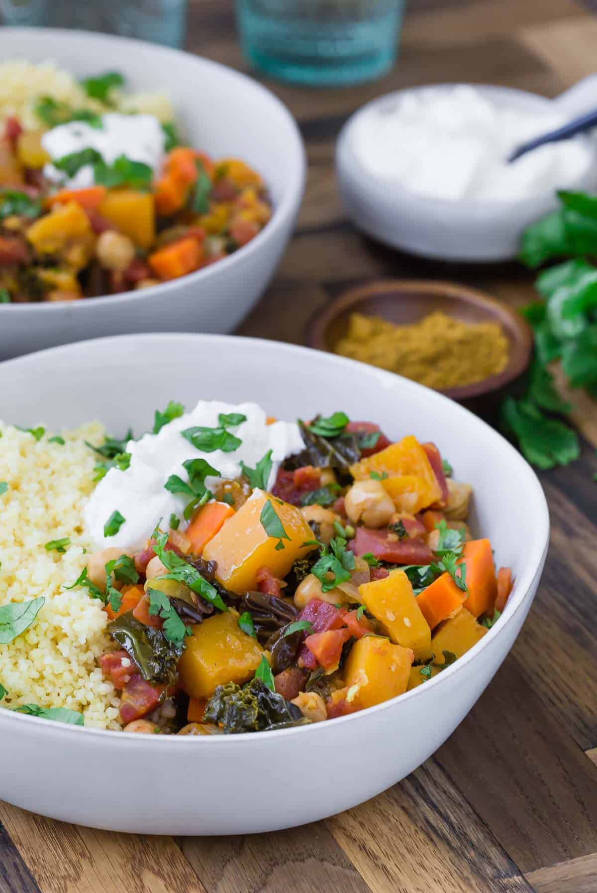 Two large white bowls with vegetarian moroccan squash stew with a small bowl of yogurt and fresh cilantro also pictured.