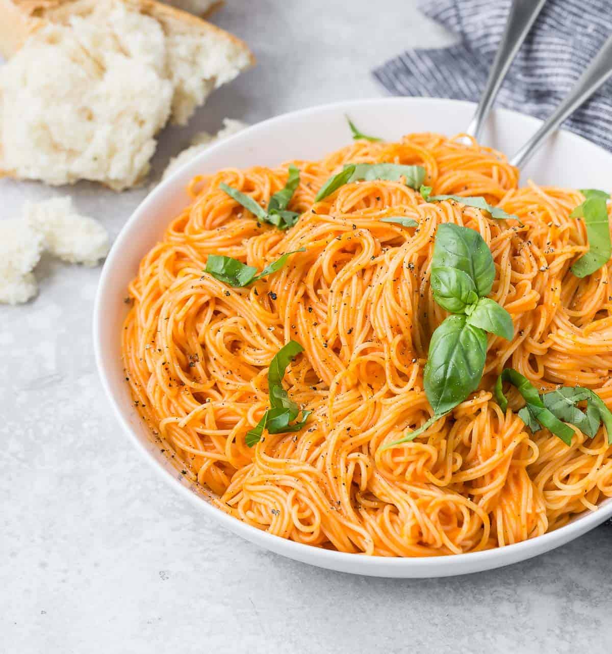 Simple pasta dish made with roasted red peppers and angel hair pasta, in a white bowl, with bread in the background.