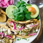 Pork tacos on a plate with cilantro, avocados, and pickled red onions.