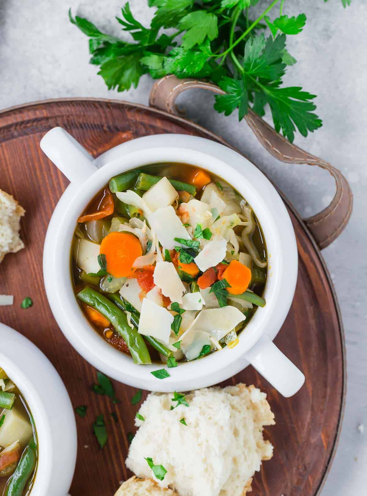 Overhead view of vegetable soup in a white bowl with handles. Bread also pictured.