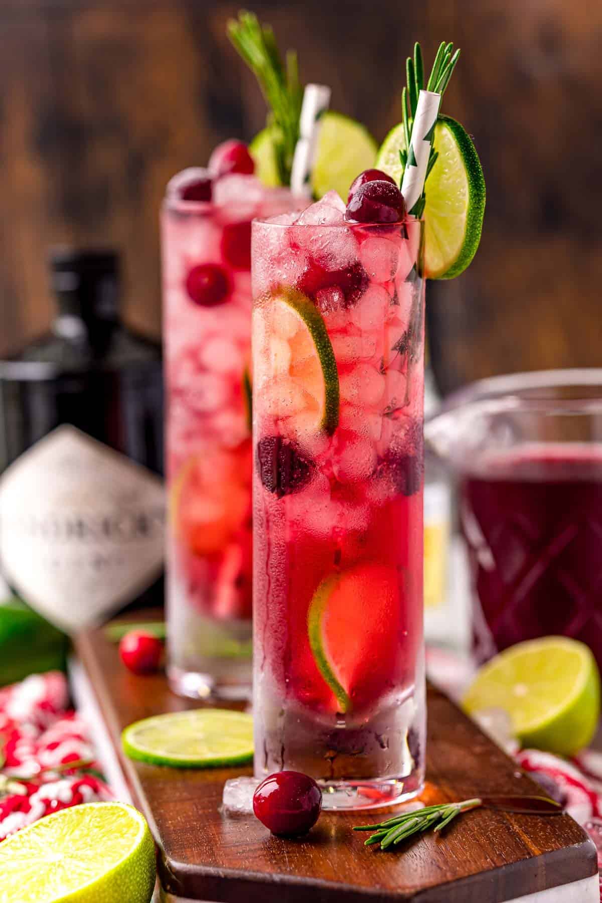 Two bright red cranberry gin and tonics with limes, fresh rosemary, and ingredients in the background.