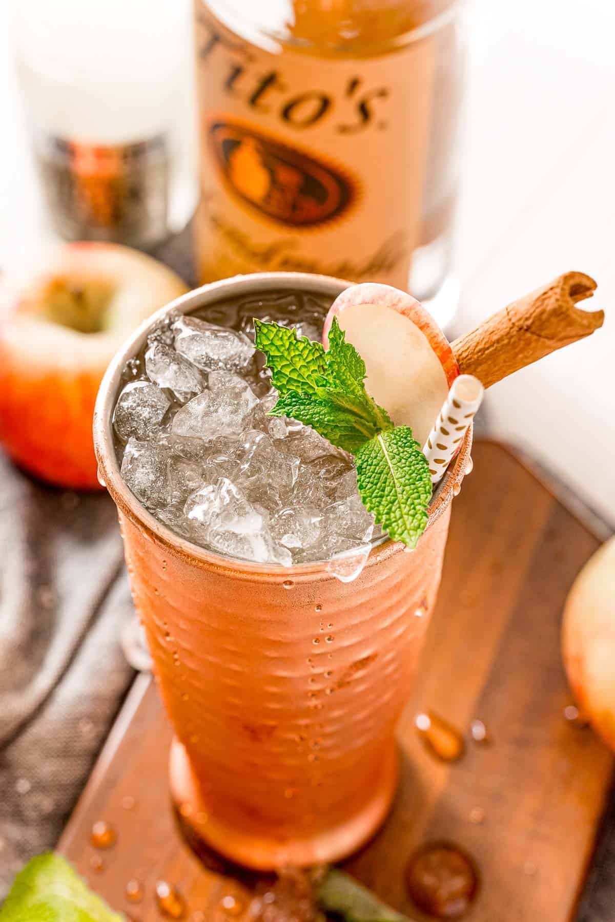 An iced drink in a copper cup, garnished with apple, mint leaves, and a cinnamon stick