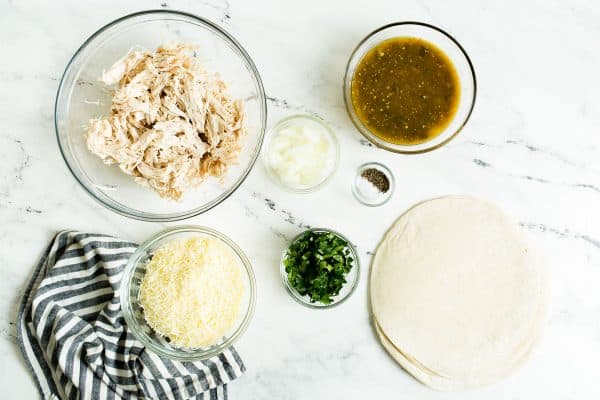 Overhead view of ingredients: tortillas, shredded chicken, salsa verde, cheese, spices.