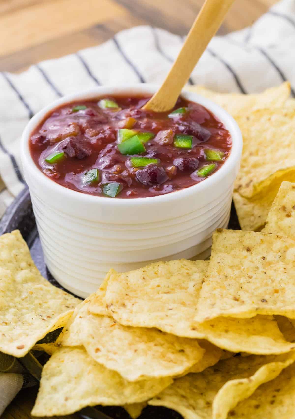 Small white bowl filled with cranberry salsa and a wooden spoon, surrounded by chips.