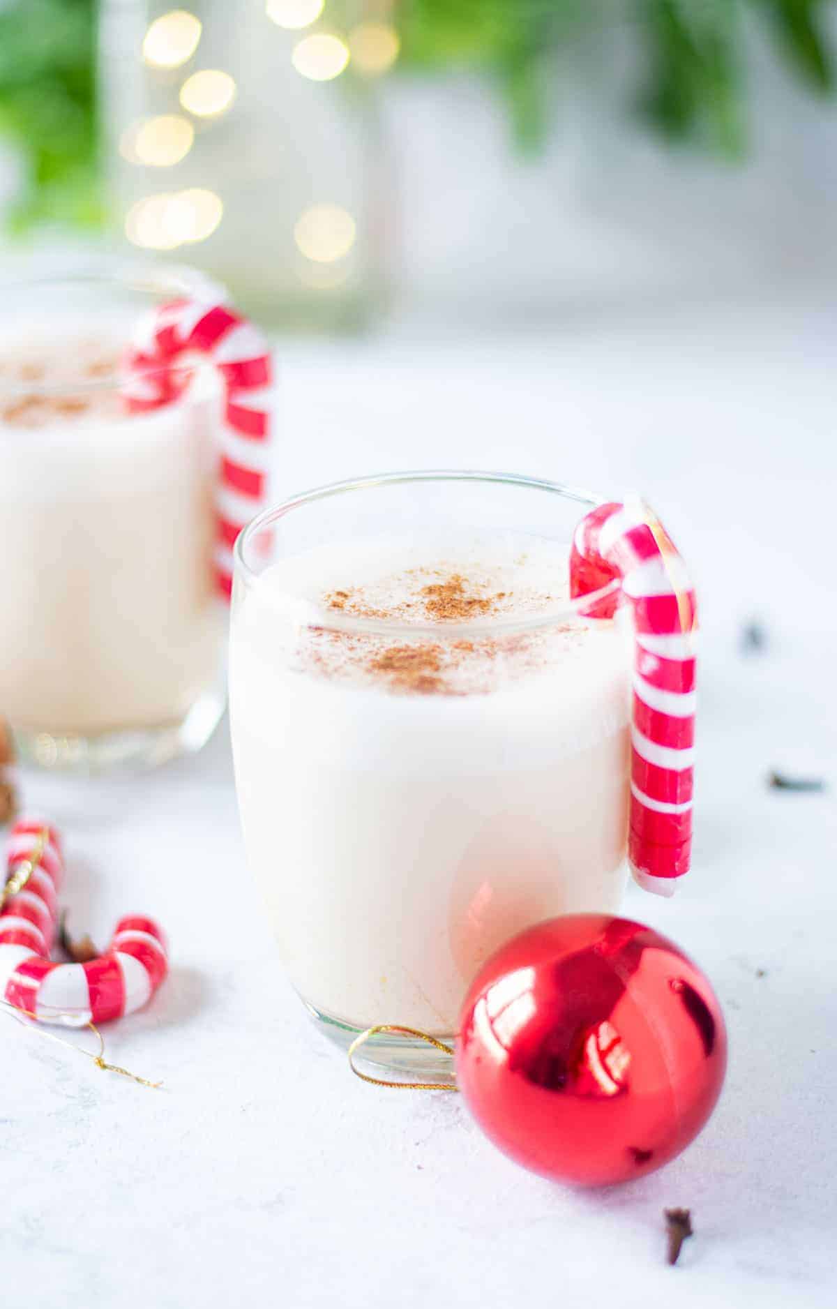 Small glass of eggnog with another one in the background. Festive Christmas ornaments scattered around.