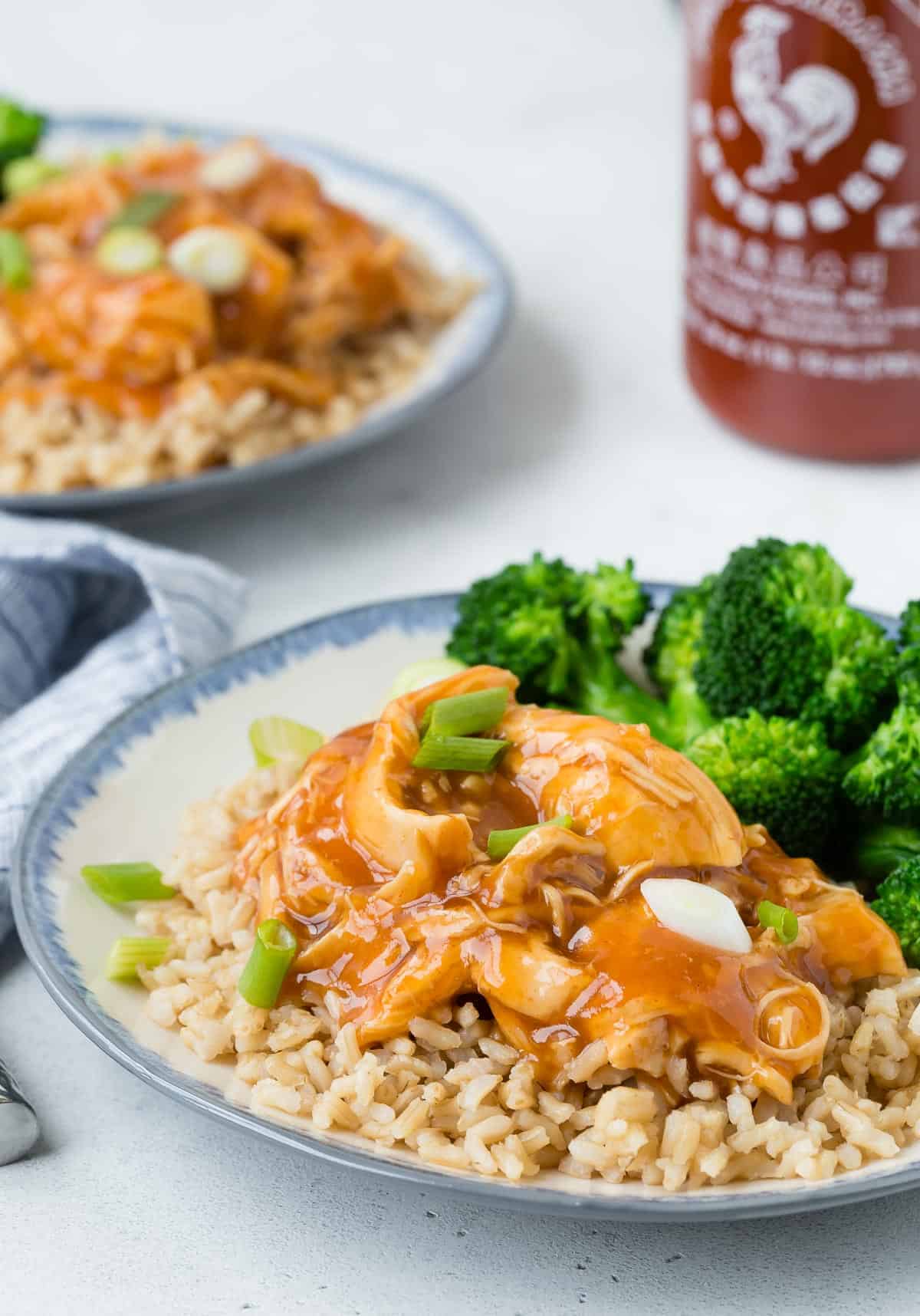 Chicken, rice, and broccoli on a plate with sriracha in the background.