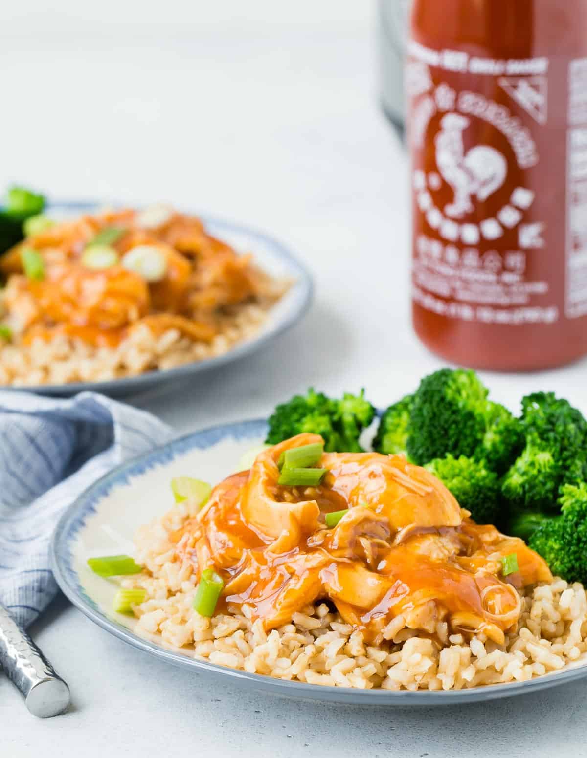 Cooked chicken, rice, and broccoli, with a bottle of sriracha in the background.