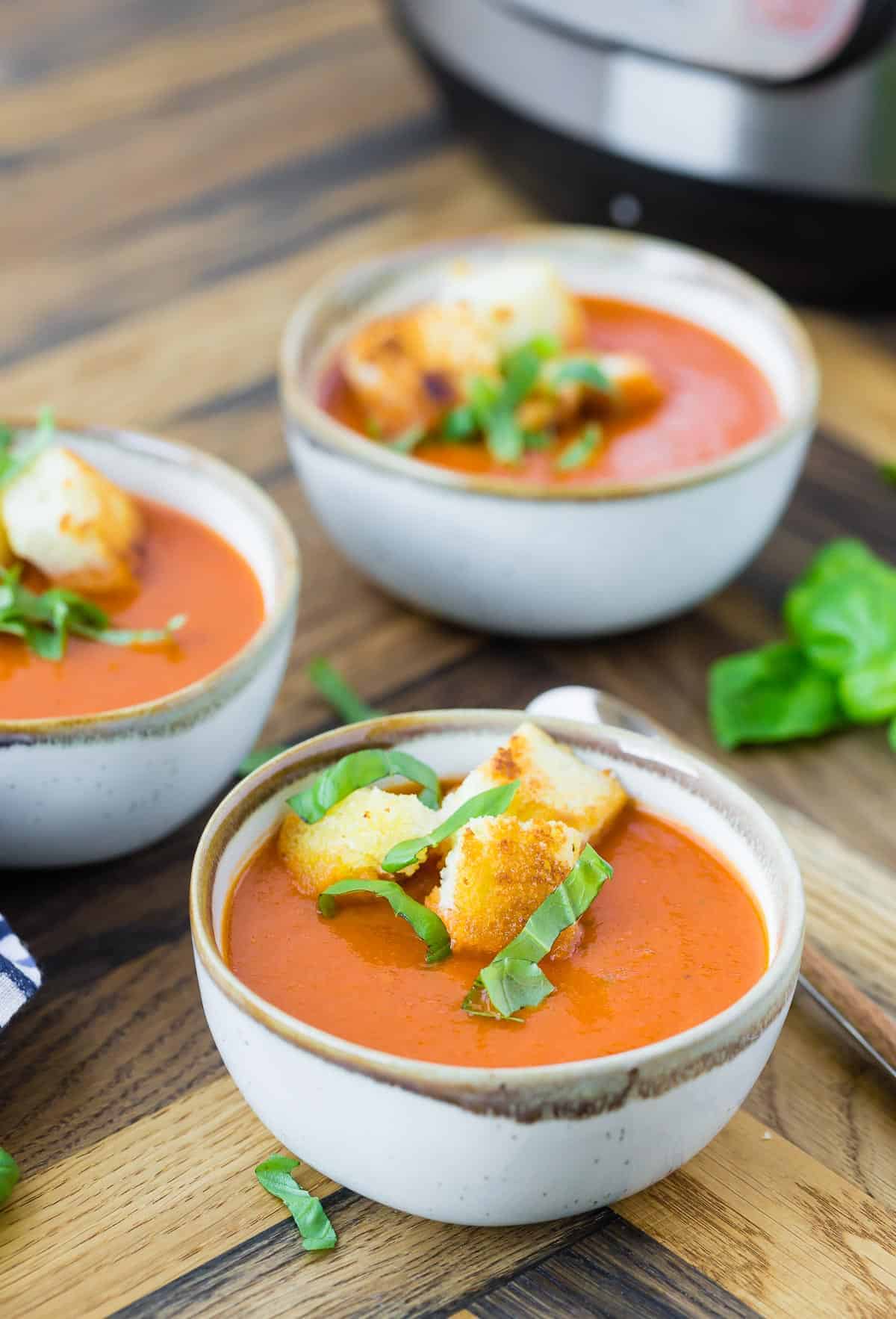 Three bowls of tomato soup on a wooden background with fresh basil.