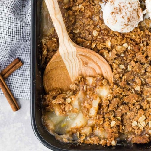 Peach crisp with ginger in a black baking dish, being scooped out with a wooden spoon.