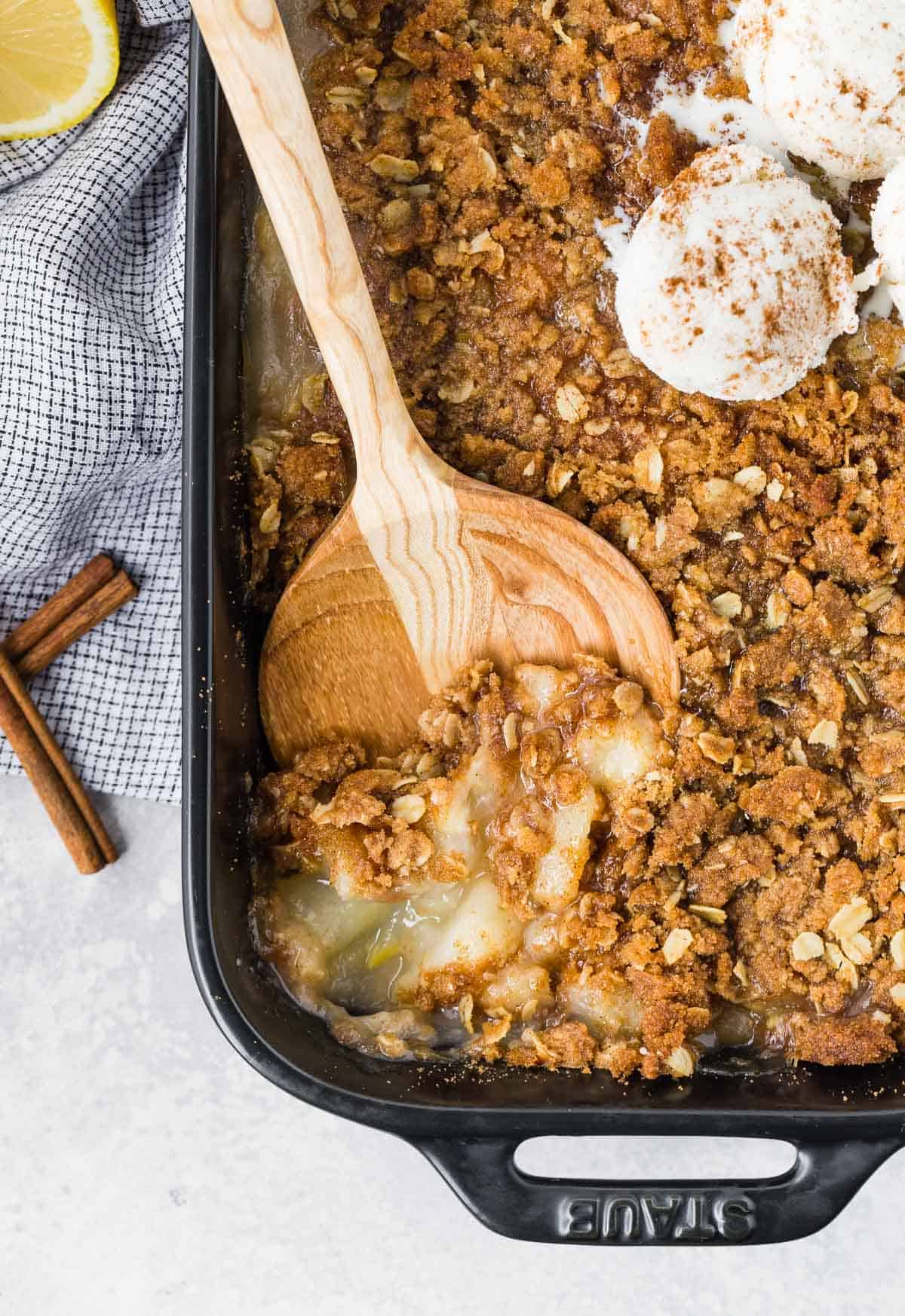 Peach crisp with ginger in a black baking dish, being scooped out with a wooden spoon.
