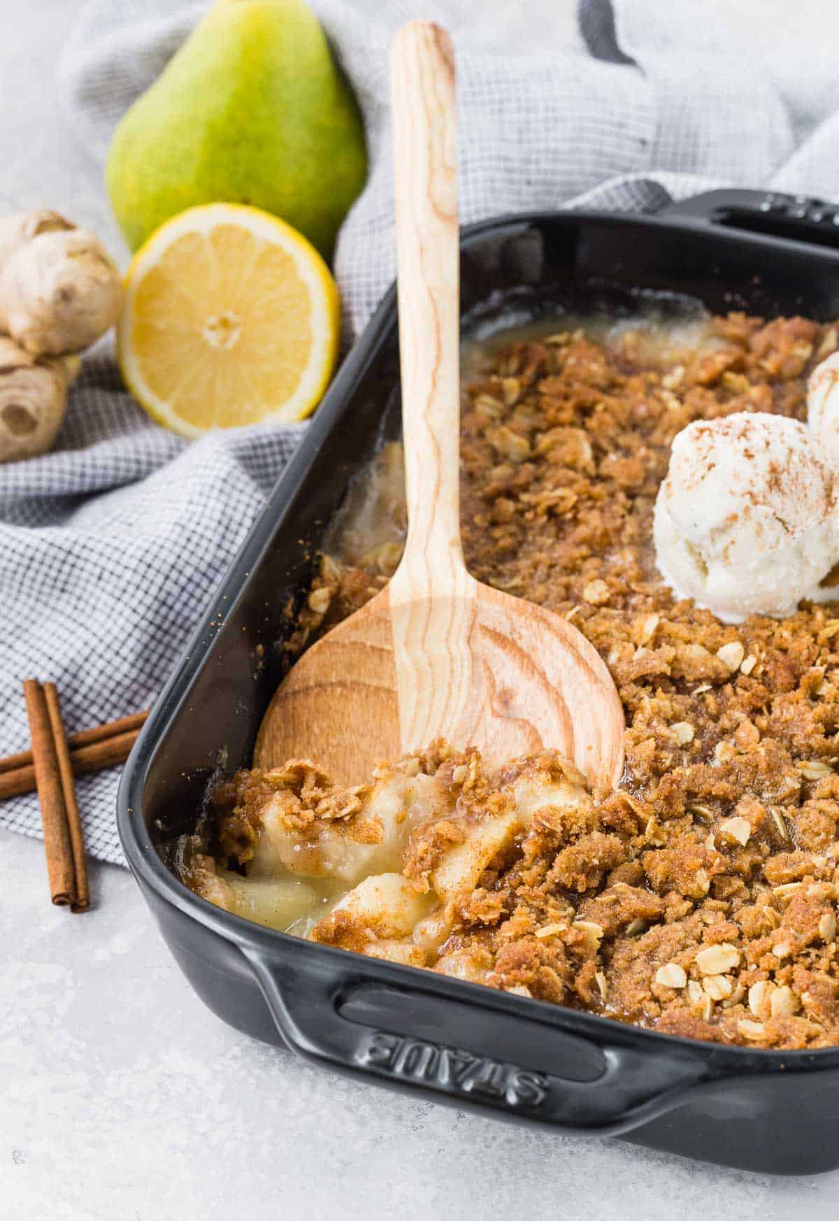 Pear and ginger crisp being scooped out of a black baking dish with a wooden spoon.