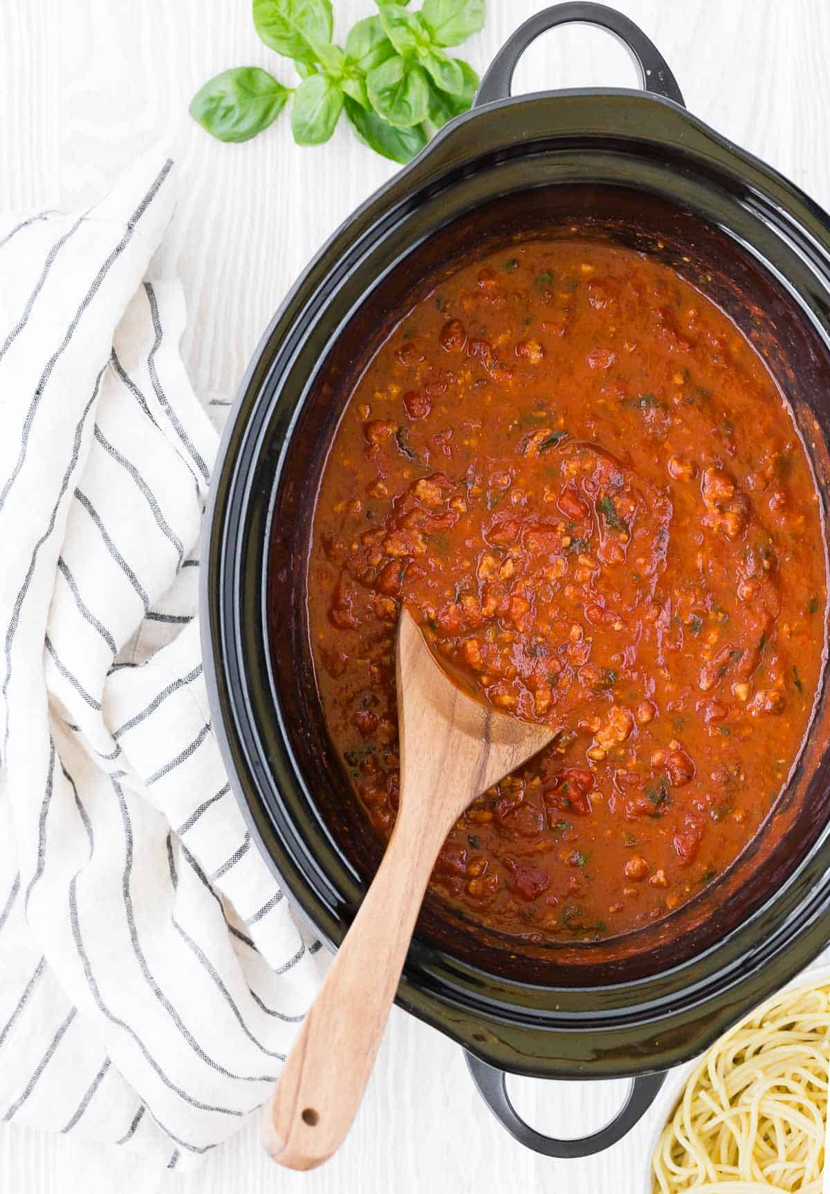 Spaghetti sauce in a black slow cooker with a wooden spoon.