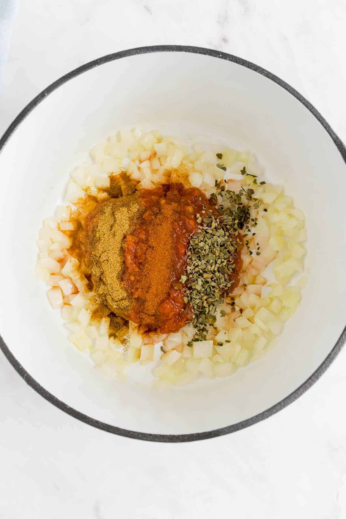 Chopped onions, ground spices, dried herbs, and tomato paste arranged in a white mixing bowl.