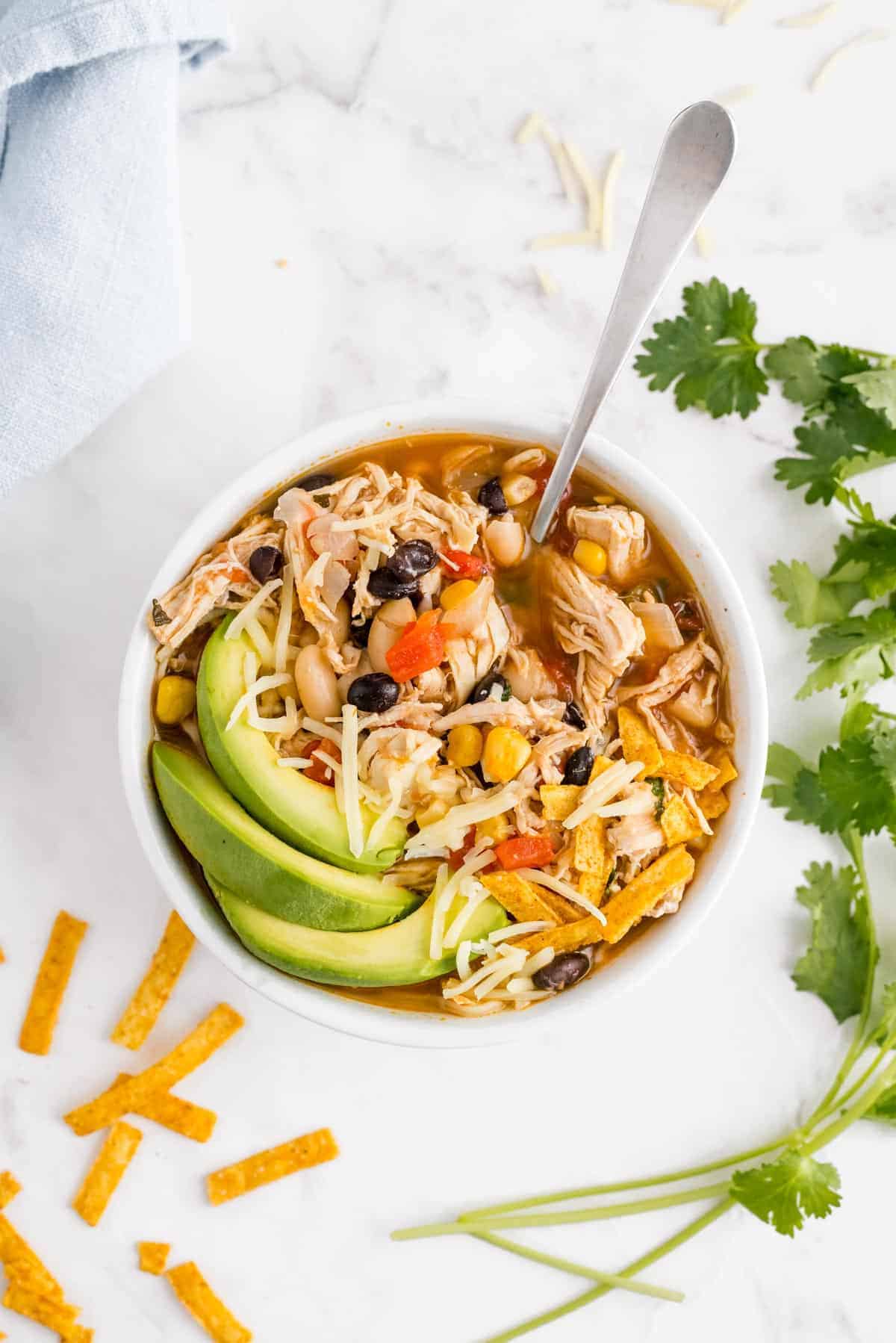 Overhead view of a bowl of chili made with chicken, with a spoon and garnishes.