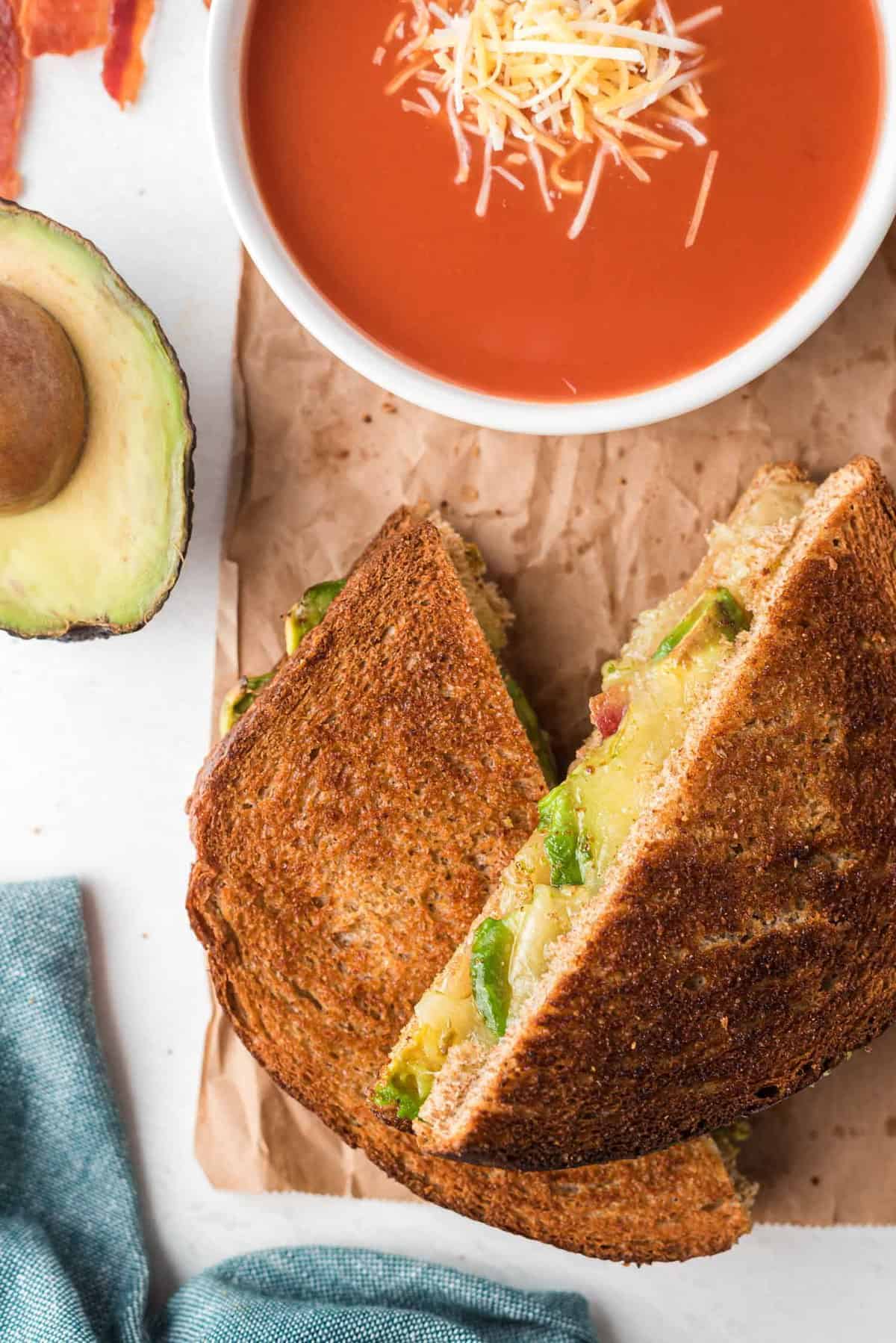 Sandwich halves that halve been toasted, filled with avocado, cheese, and bacon, served with tomato soup.