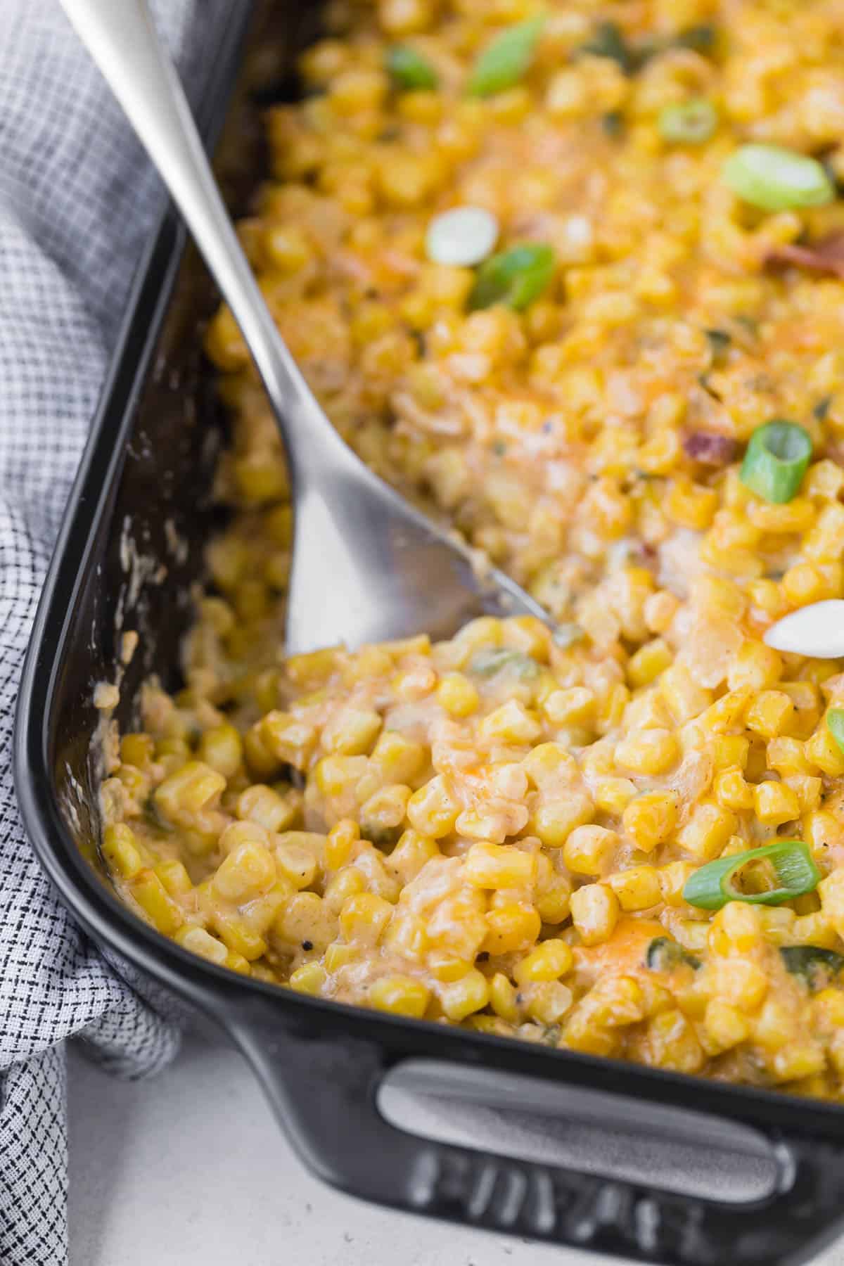 Cheesy corn casserole in a black casserole dish with a silver spoon.