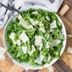 Overhead view of a white bowl with an arugula salad with parmesan and lemon on a wooden background.