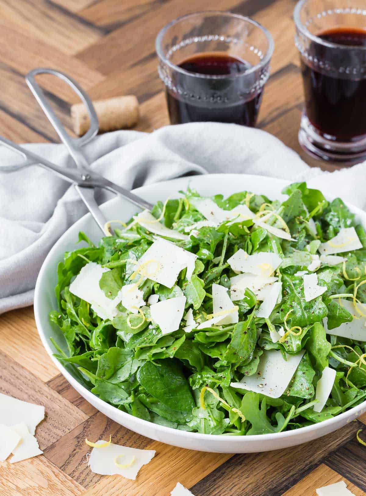 Arugula, parmesan and lemon zest in a white bowl on a wooden background, with two glasses of red wine.