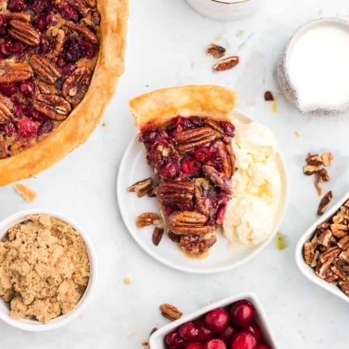 Overhead view of cranberry pecan pie on a white plate surrounded by ingredients and more pie.