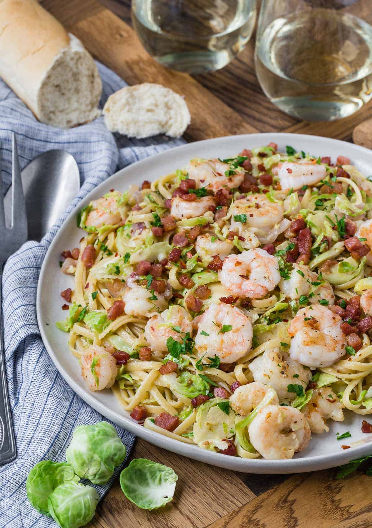 Large plate of pasta with shrimp, bread and wine in background, fresh brussels sprouts in foreground.