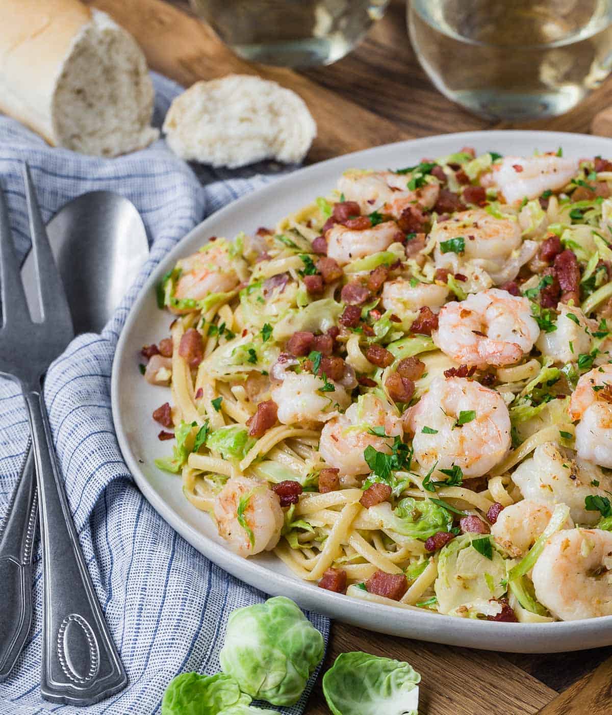 A large serving plate of pasta with brussels sprouts and shrimp. Bread and wine in background.