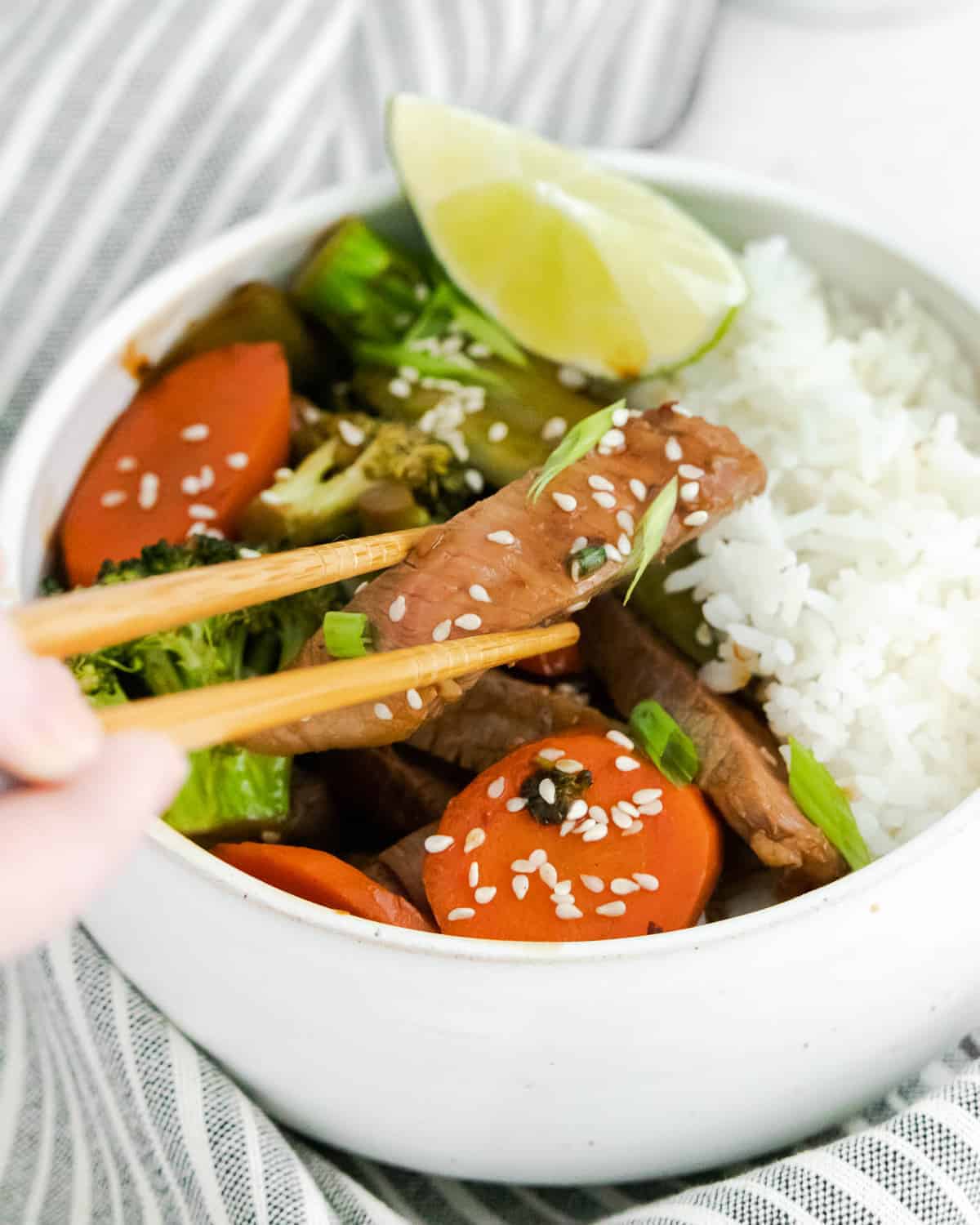 Chopsticks taking a piece of cooked steak out of a bowl of rice, beef, and vegetables.