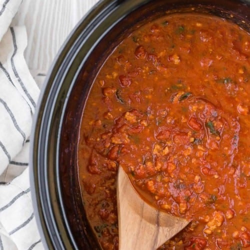 A wooden spoon inserted into a black crockpot full of spaghetti sauce.