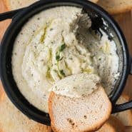 Dip in a black pan with a small pieces of crisp bread. Text overlay reads "baked goat cheese dip with lemon and thyme, rachelcooks.com"