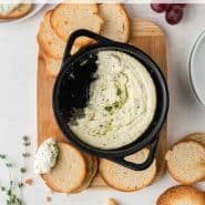Dip in a black pan with a small pieces of crisp bread. Text overlay reads "baked goat cheese dip with lemon and thyme, rachelcooks.com"